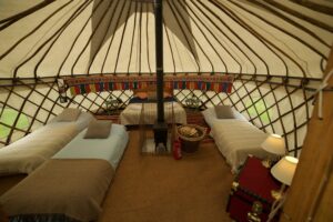 Interior of a furnished yurt with several beds, a central stove, and lamps.