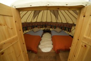 Inside a yurt with two single beds, pillows, and a central rug seen through open wooden doors.
